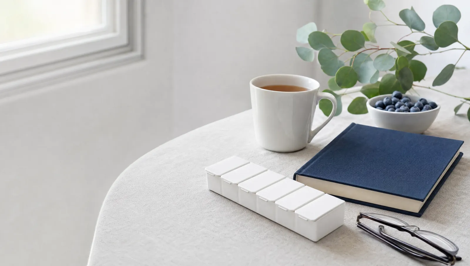 Breakfast table with tea, blueberries, a notebook, reading glasses and a pill organizer