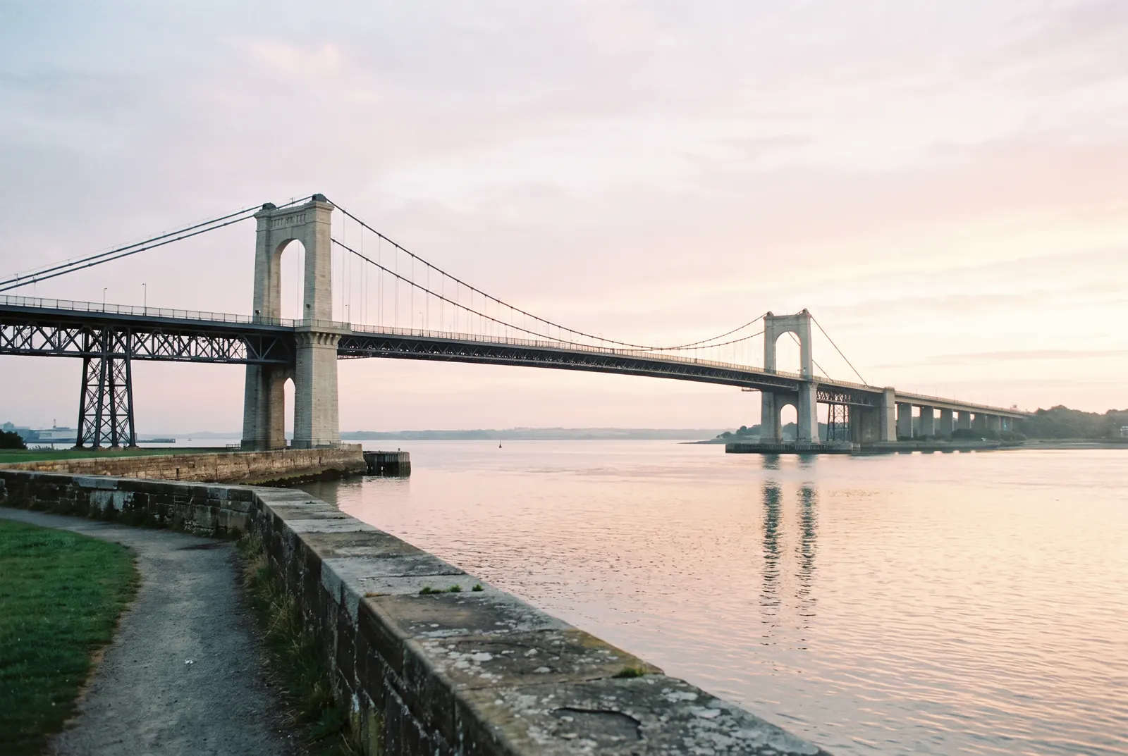 Suspension bridge over a harbor entrance at sunrise, seen from a waterfront path.
