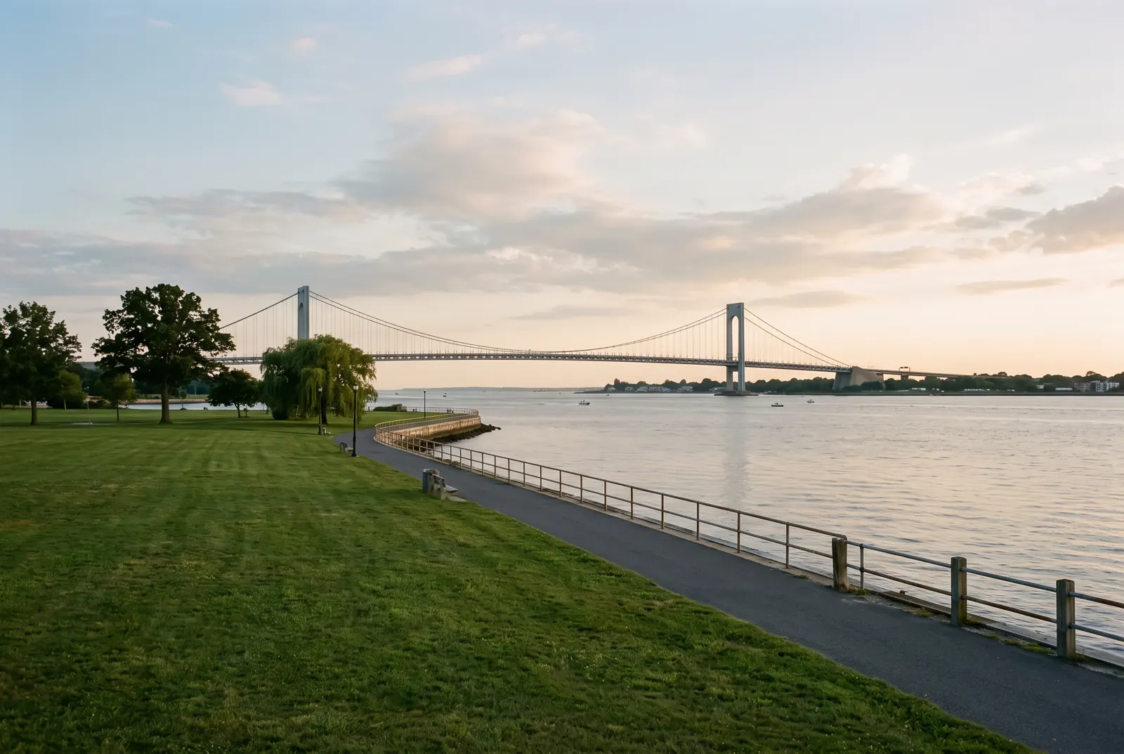 A waterfront path beside a calm bay with a suspension bridge in the distance at sunset