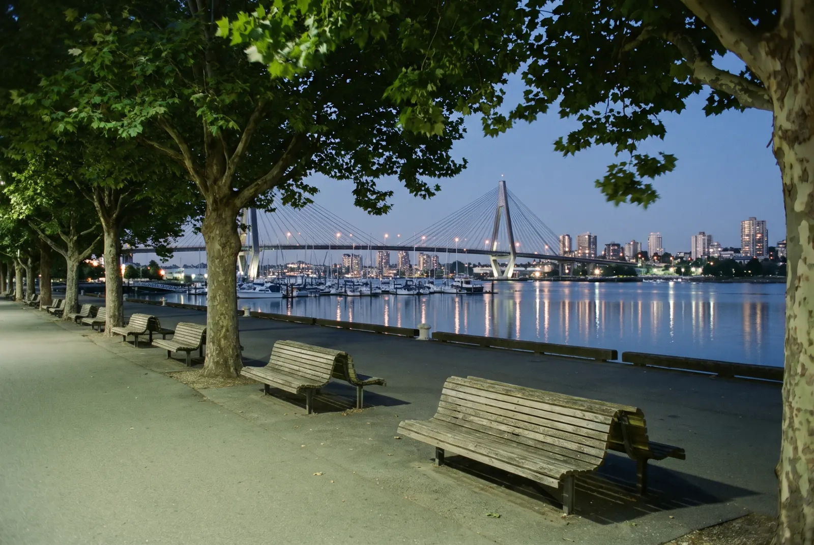 Riverside promenade with benches and trees overlooking a harbor, a bridge and a skyline at dusk.