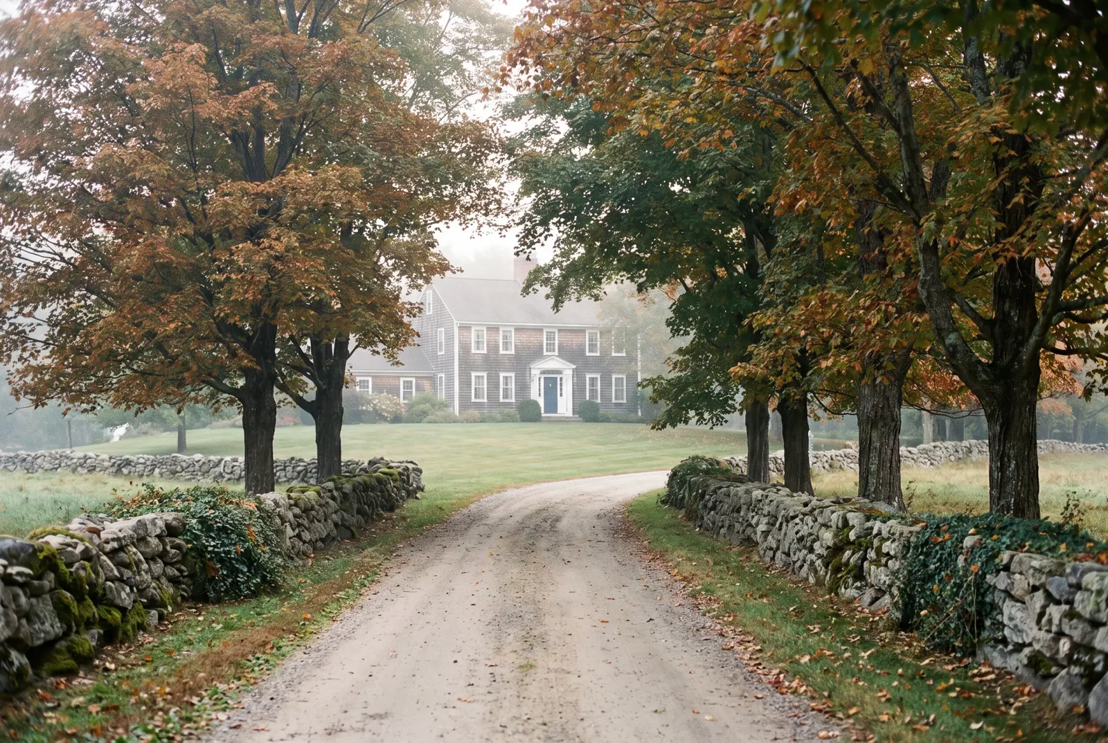 Country road lined with stone walls and autumn maples, with a colonial house set back on a broad lawn.