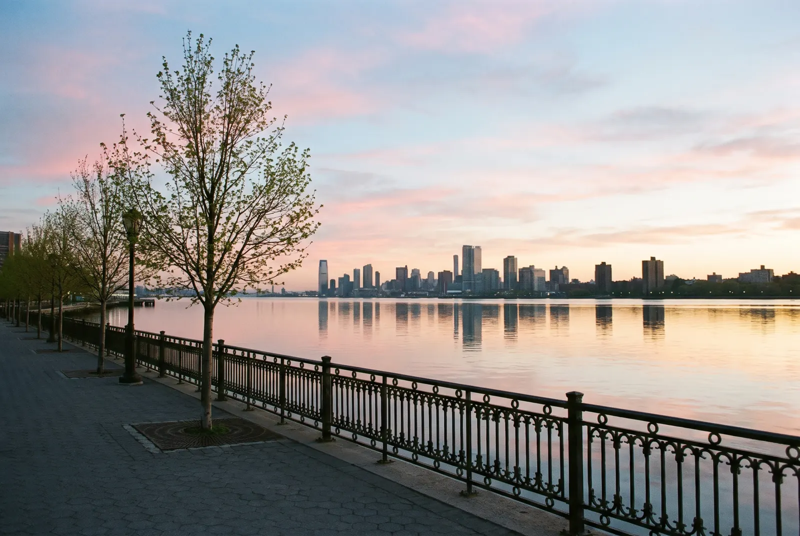 A riverside walkway at sunrise with a city skyline across the water
