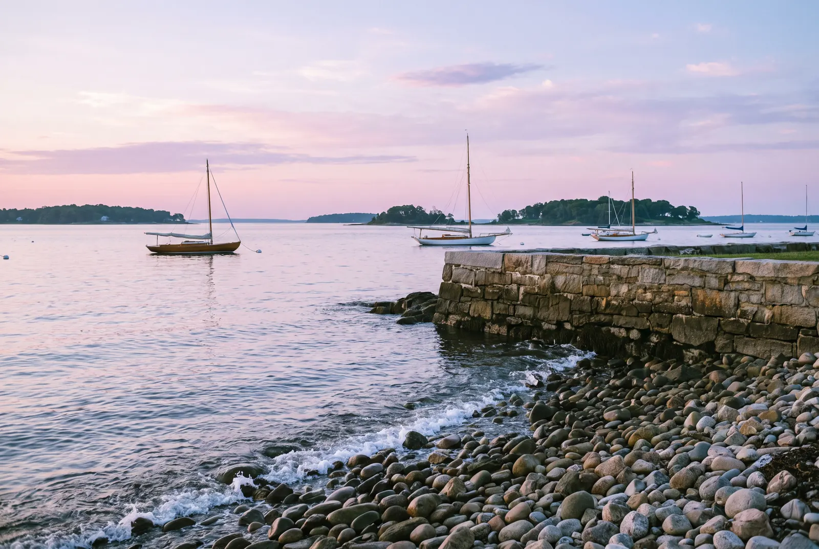 A rocky shoreline at dawn with sailboats moored offshore and wooded islands in the distance