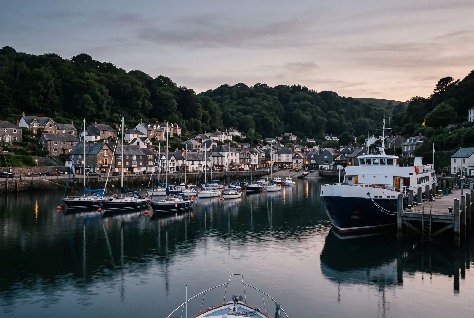 Harbor village at dusk with wooded hills behind rooftops, moored sailboats and a docked ferry
