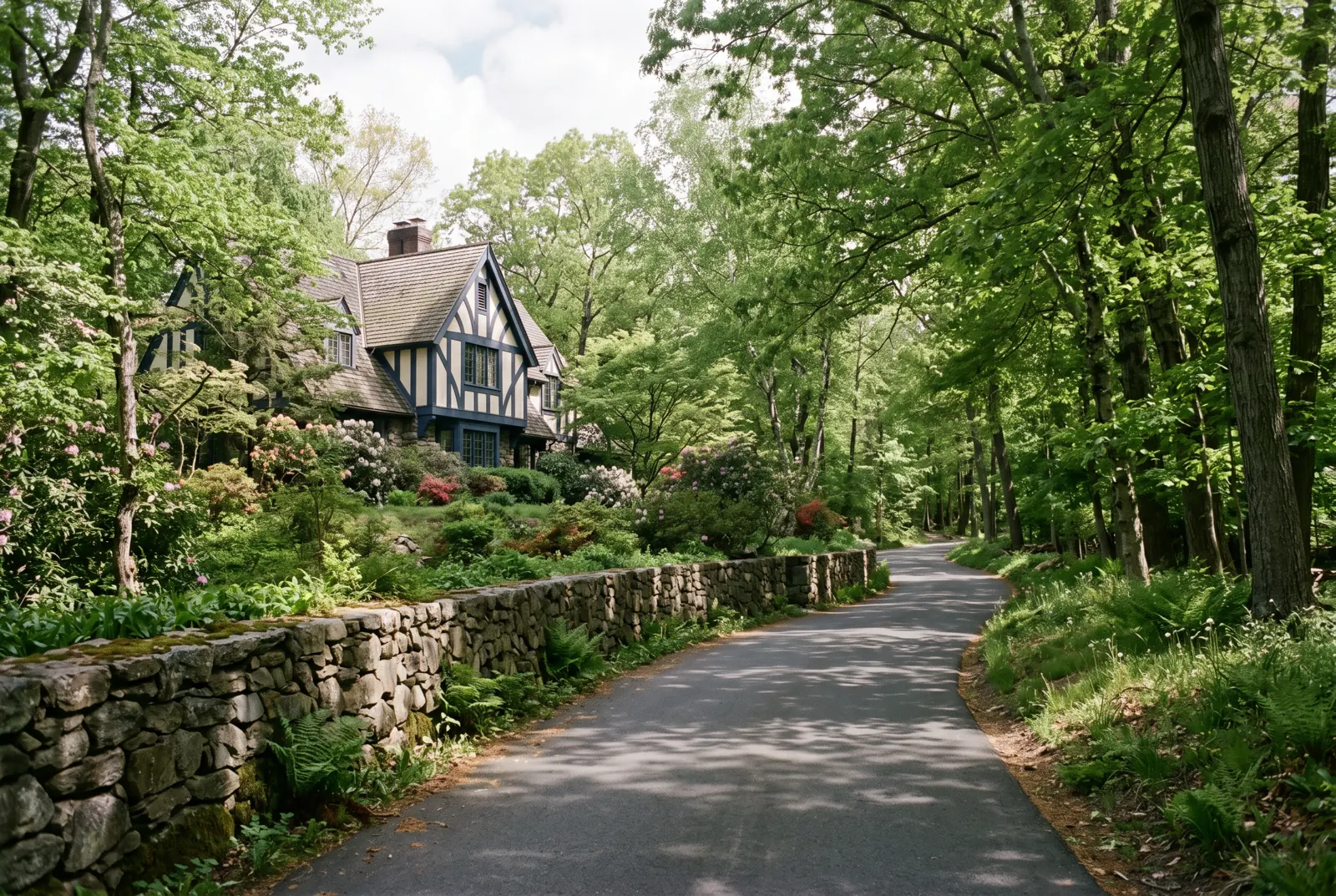 A winding wooded road with a stone wall and a Tudor-style house glimpsed through the trees