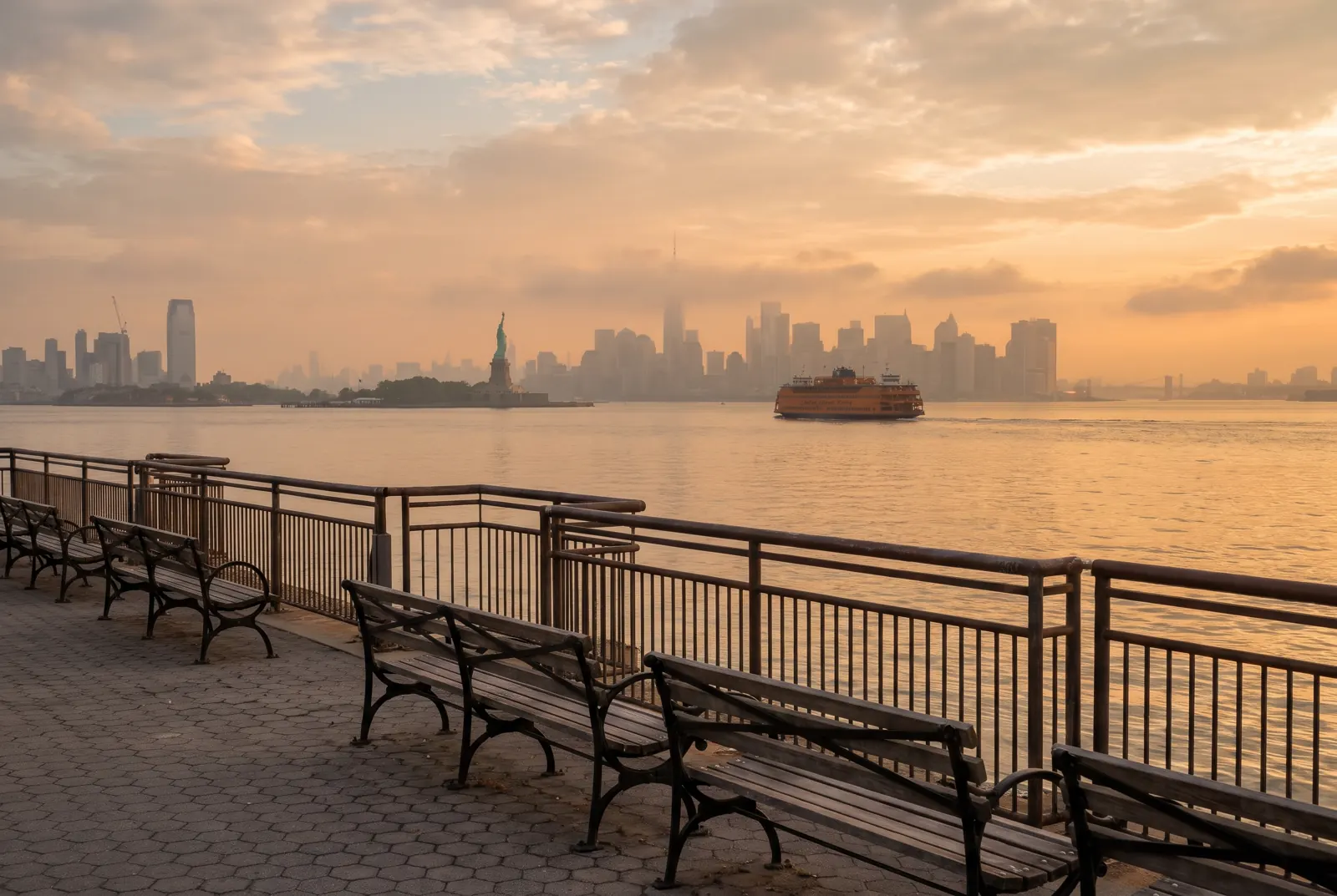 A ferry crossing the harbor seen from a waterfront promenade, with a distant city skyline in the haze