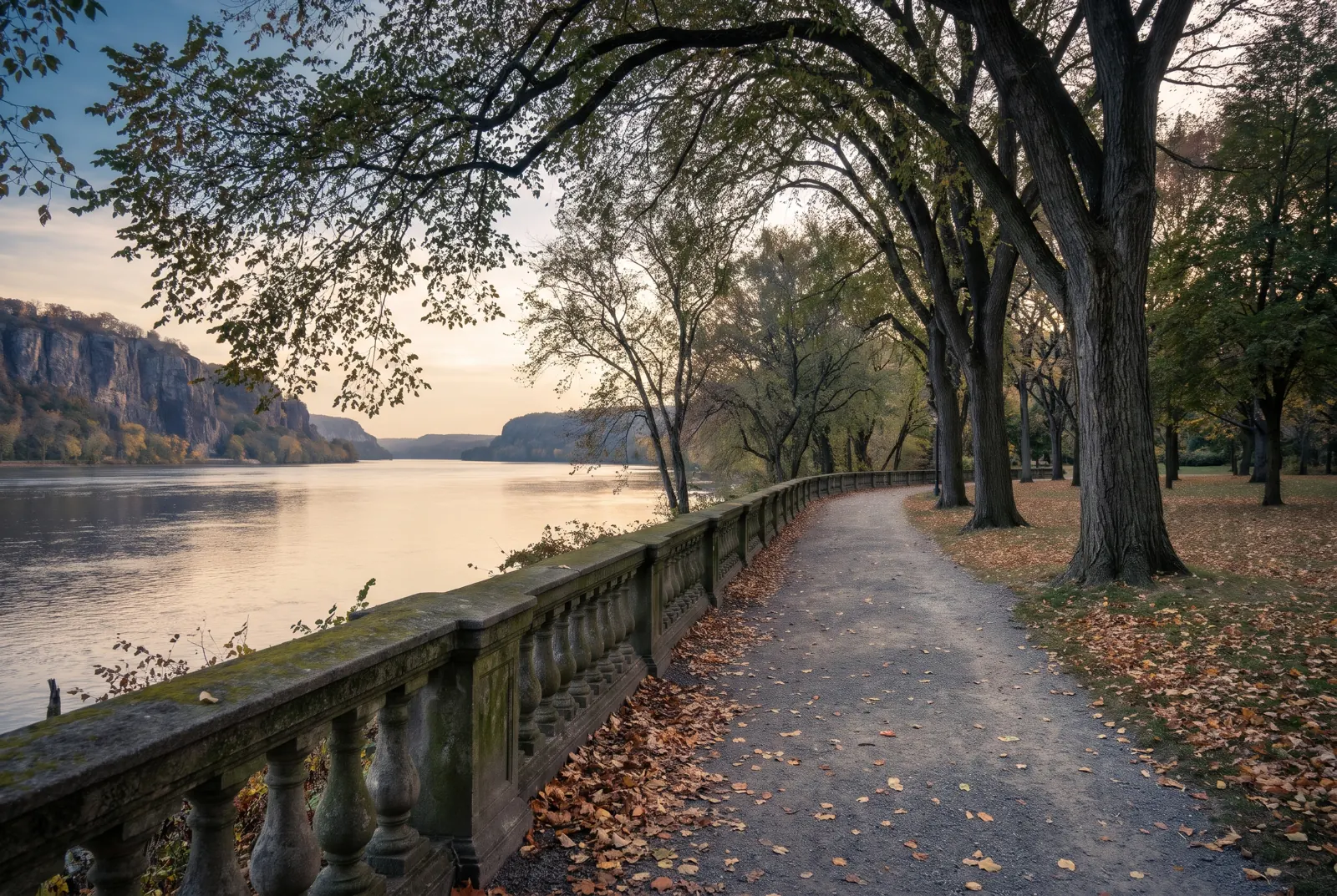 Park path with a stone balustrade and tall trees beside a wide river at golden hour.