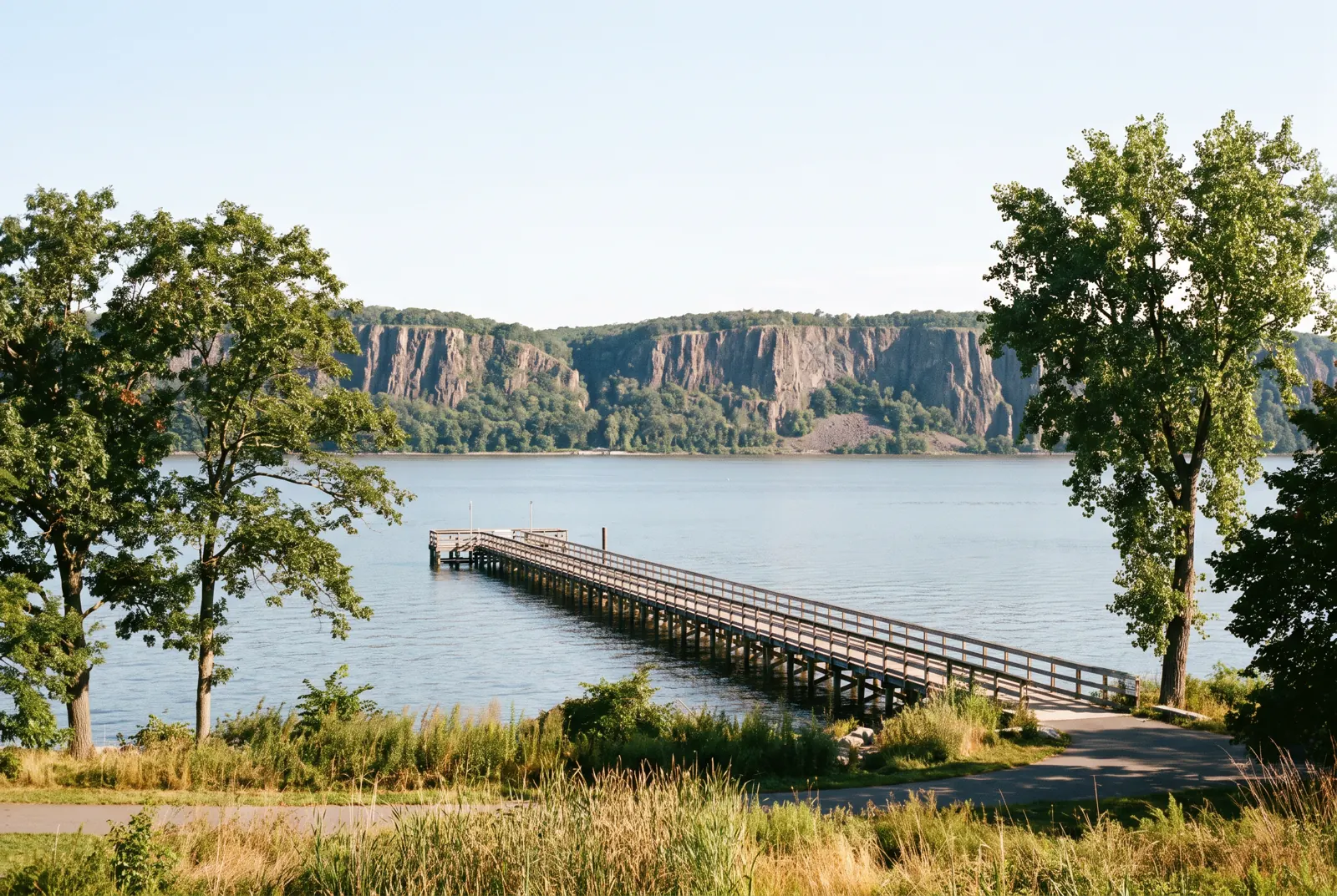 A long pier reaching into a wide river on a clear morning, with tall cliffs on the far shore